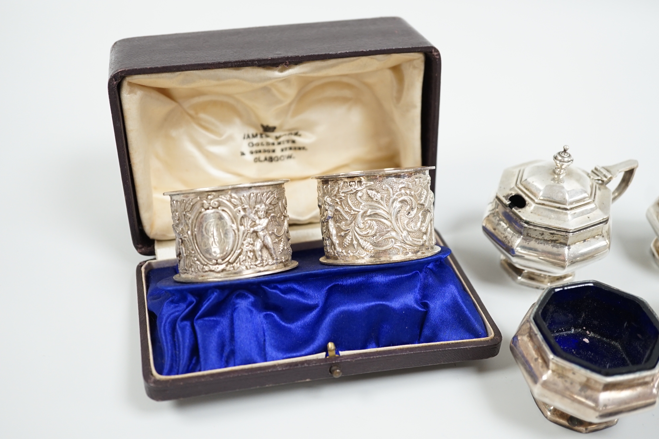 A cased pair of Edwardian embossed silver napkin rings, Birmingham, 1904, a five piece silver condiment set, Birmingham, 1926, two plated condiments and a match sleeve.